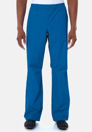 Vorderansicht einer Person, die blaue wasserdichte Hosen und schwarze Schuhe trägt, mit entspannten Armen seitlich am Körper stehend vor weißem Hintergrund.