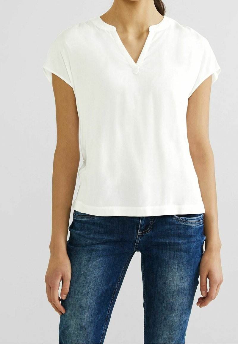 White short-sleeve blouse with a V-neck, soft texture, and a relaxed fit. Worn with dark blue denim jeans featuring slight fading.