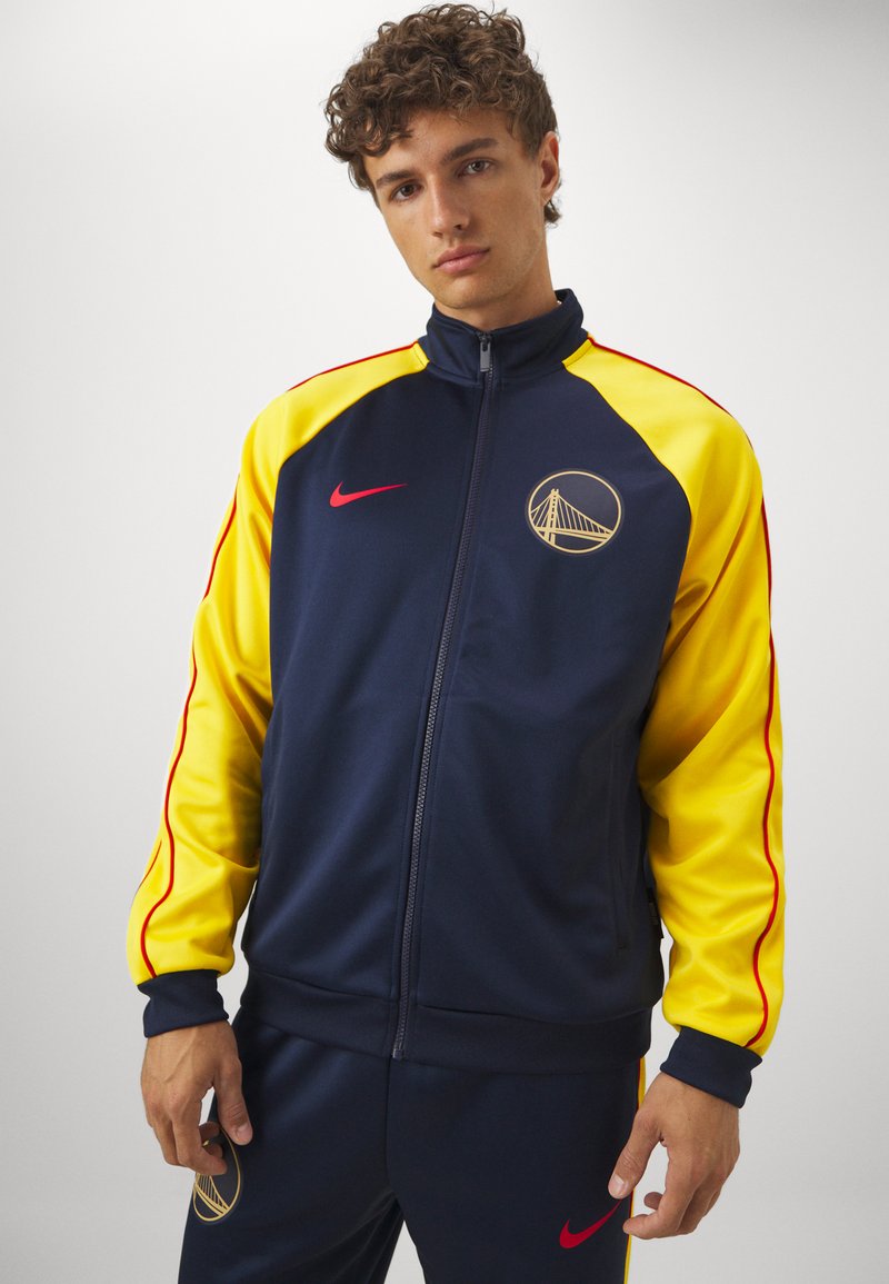 Giacca da track blu navy con maniche gialle, dettagli rossi e un logo dorato. Realizzata in materiale liscio con zip frontale e polsini elasticizzati.