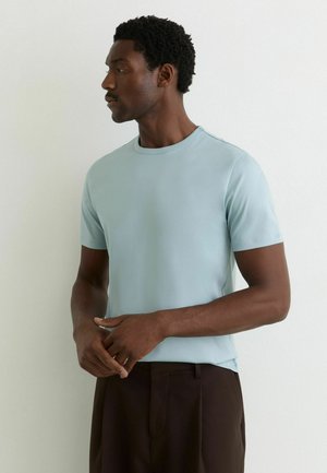 Mand med kort krøllet hår iført en lyseblå kortærmet skjorte og mørke bukser, der kigger til siden med hænderne foldet.