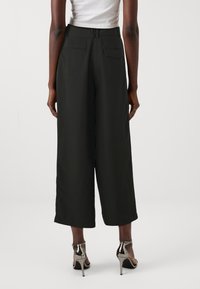 Pantaloni neri a gamba larga con una texture liscia, caratterizzati da una vita alta, tasche posteriori e una vestibilità rilassata, abbinati a scarpe con tacco alto argentate.