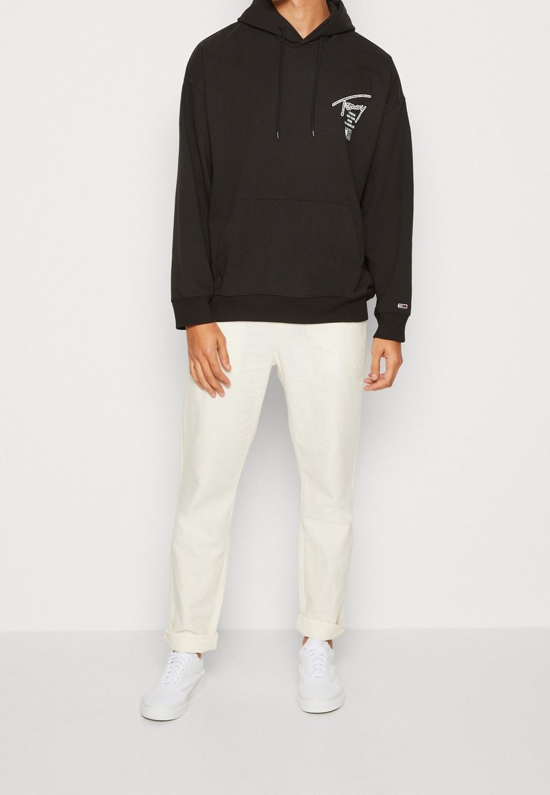 Personne portant un sweat à capuche noir avec un texte blanc sur la poitrine, un pantalon écru à revers et des baskets blanches, debout devant un fond clair uni.
