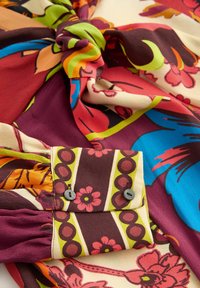 Blusa floreale multicolore con dettaglio a nodo; presenta stampe vivaci nelle tonalità di bordeaux, blu, arancione e verde; polsini con bottoni.