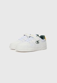 Baskets blanches en cuir lisse, avec des accents en maille, une languette de talon verte, des détails jaunes et un laçage sécurisé avec une sangle en Velcro.