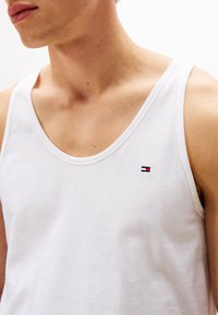 Nahaufnahme einer Person, die ein weißes, ärmelloses Tanktop trägt, mit einem kleinen gestickten rechteckigen Logo in Rot, Weiß und Blau auf der Brust.
