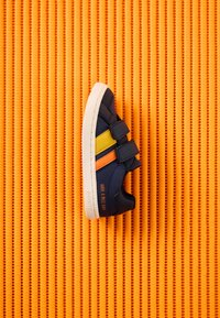 Zapatilla deportiva azul marino con suela blanca, rayas amarillas y naranjas, dos tiras de velcro y texto "¡Que tengas un buen día!" sobre un fondo texturizado naranja.