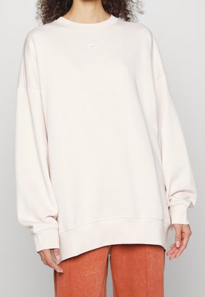 Personne portant un sweat-shirt Nike ample de couleur beige clair à manches longues et un pantalon orange brûlé, debout devant un fond uni.