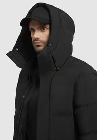 Sort pufferjakke med høj krave og oversize hætte, der har en robust lynlås og justerbare snore. Glat mat finish og firkantet form.