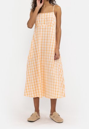 Robe à carreaux orange et blanche avec bretelles spaghetti, encolure carrée et détail à boutons. Le mannequin porte des chaussures beige sans lacets.