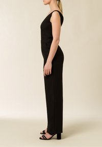 Svart byxdress med en åtsittande livdel och breda axelremmar, med en definierad midja och raka ben, kombinerad med svarta högklackade sandaler.