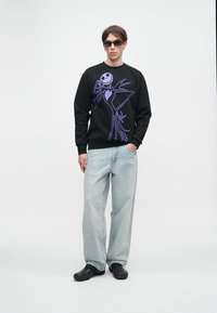 Jovem vestindo um moletom preto com um design de esqueleto estilizado roxo, jeans azul claro, sapatos pretos e óculos de sol escuros, em pé sobre um fundo branco.