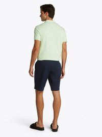Camisa polo verde menta com gola, combinada com calções azul-marinho. O modelo usa sandálias deslizantes pretas. Vista de trás.