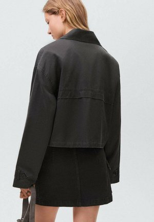 Veste noire courte avec une texture lisse et un col, assortie à une jupe mini noire. Les deux présentent un design minimaliste.