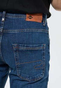 Jean en denim bleu à coupe décontractée, avec une poche arrière visible et des surpiqûres orange contrastantes. Étiquette en cuir avec le nom de la marque sur la taille.
