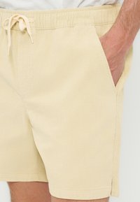 Μπεζ σορτς με κορδόνι στη μέση, ελαστική μέση και πλαϊνές τσέπες, φορεμένα με το ένα χέρι άνετα στην τσέπη.