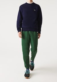 Felpa navy con polsini a coste, abbinata a pantaloni della tuta verdi. Sneakers nere multicolori con lacci turchesi. Fit semplice e rilassato.