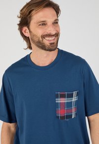Camiseta de algodón en azul oscuro con mangas cortas, que presenta un bolsillo de parche a cuadros en rojo, negro y blanco. Textura suave y diseño casual.