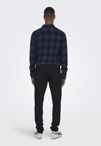 Chemise à carreaux noire et bleue à boutons avec des manches longues, assortie à un pantalon noir et des baskets blanches. Fond uni.