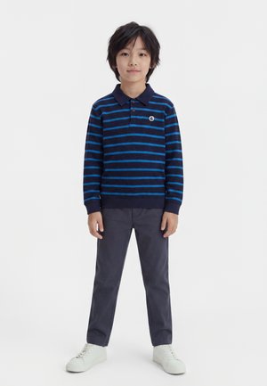 Garçon debout portant un polo à manches longues rayé bleu marine et bleu, pantalon gris et baskets blanches sur un fond blanc uni.