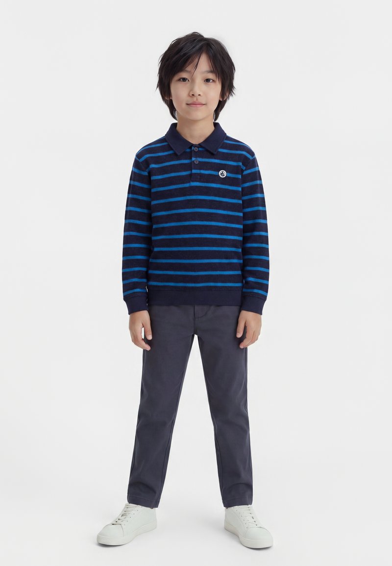 Garçon debout portant un polo à manches longues rayé bleu marine et bleu, pantalon gris et baskets blanches sur un fond blanc uni.