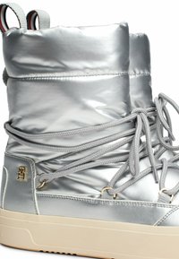 Metallic zilveren laars met een gevoerde bovenkant, voorzien van grijze veters, gouden hardware-accenten en een gestructureerde rubberen zool.