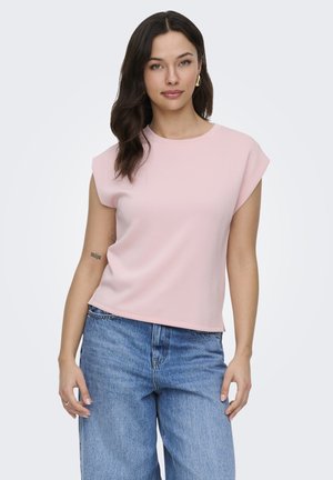 Kvinde med langt mørkt hår iført en lyserød ærmeløs top og blå jeans, stående mod en ensfarvet lys baggrund.