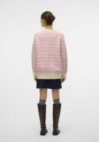 Femme avec un chignon portant un pull à motifs "PARIS" rose et beige, une jupe en denim, des chaussettes grises et des bottes marron montantes, vue de dos.