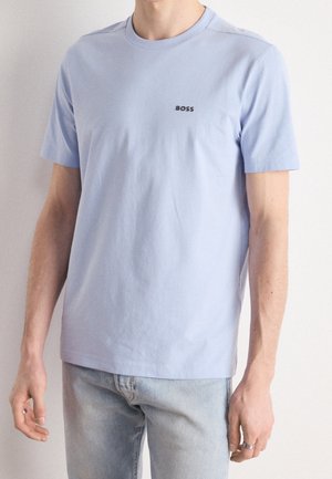 Homme portant un t-shirt bleu clair à manches courtes avec le logo "BOSS" sur la poitrine et un jean bleu clair, debout devant un fond uni.
