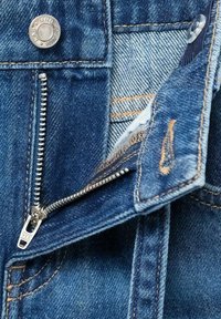 Lähellä denim-takkia, jossa on metallivetoketju, hopeanvärinen liukusäädin, messinkinappia ja erottuva ompelu. Kankaan rakenne on näkyvissä.