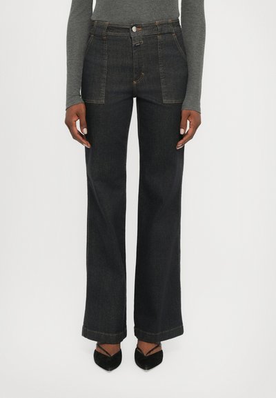 Jeans en denim foncé à jambes larges et taille haute, avec des coutures contrastées et des poches avant. Le mannequin les associe à des talons noirs à bout pointu.