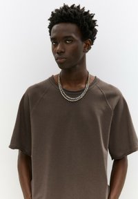 Barna, oversized fazonú póló, lekerekített nyakkal és raglan ujjal. Texturált anyaggal és rétegezett ezüst lánc nyakláncokkal rendelkezik.