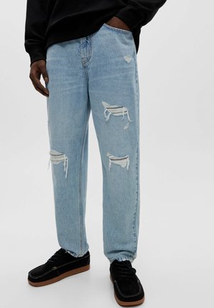 Mann trägt hellblaue, zerrissene Jeans mit ausgefransten Löchern und schwarze Sneakers mit braunen Sohlen, steht mit einer Hand in der Tasche.