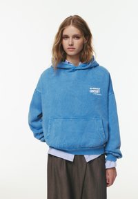 Felpa blu con cappuccio in tessuto morbido e testurizzato, dotata di tasca a marsupio e logo stampato bianco. Abbinata a una camicia a righe blu sotto.