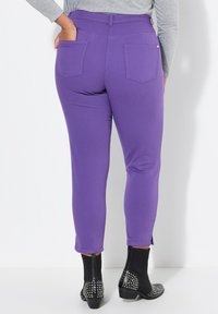 Lila Stretch-Jeans mit zwei Gesäßtaschen, figurbetontem Design und verkürzter Länge, kombiniert mit schwarzen Knöchelstiefeln mit Nietenakzenten.