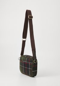 Small rectangular crossbody bag with dark green, red, and white plaid fabric and a wide adjustable brown strap hanging against a white background.
