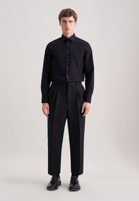 Zwart shirt met lange mouwen, hoge kraag, nauwsluitend, knoopsluiting aan de voorkant; gecombineerd met op maat gemaakte zwarte broek met plooien en zwarte schoenen.