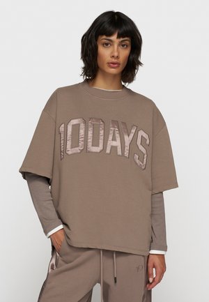 Brązowy oversize t-shirt z napisem "10DAYS" w satynowym wykończeniu. Krótkie rękawy, okrągły dekolt, noszony na szarym topie z długim rękawem. Gładka faktura materiału.