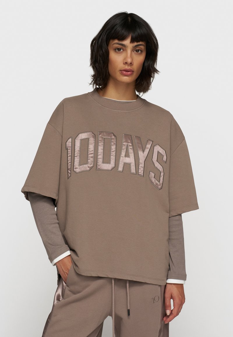 Brązowy oversize t-shirt z napisem "10DAYS" w satynowym wykończeniu. Krótkie rękawy, okrągły dekolt, noszony na szarym topie z długim rękawem. Gładka faktura materiału.