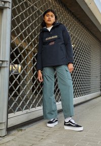 Námorní pulovrová bunda s logem a vlajkou, v kombinaci s světle zelenými cargo kalhotami a černými teniskami se bílou podrážkou, stojící na dlažebním povrchu.