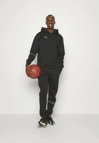 Čierna mikina s kapucňou a tepláky so zúbkovanými akcentmi, drží basketbal. Tkanina vyzerá jemne, športové topánky majú podrobný dizajn.