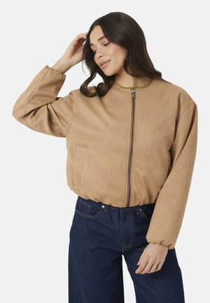 Vrouw met lang donker haar, gekleed in een lichtbruine jas met rits en donkerblauwe jeans, poseren met één hand die haar haar aanraakt.