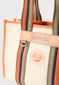 Υφασμάτινη τσάντα χειρός σε μπεζ με πορτοκαλί δερμάτινες λεπτομέρειες, με ριγέ χερούλια σε πορτοκαλί, καφέ και ναυτικό μπλε. Κονκάρδα με το λογότυπο "ROLAND GARROS" στο μπροστινό μέρος.