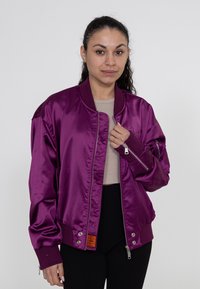 Paarse satijnen bomberjack met ribgebreide manchetten, een rits aan de voorkant en een oranje label. Het heeft een relaxed fit en zijzakken.