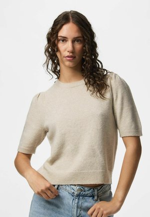 Femme aux cheveux bruns bouclés portant un pull beige à manches courtes et un jean bleu clair, posant devant un fond uni.