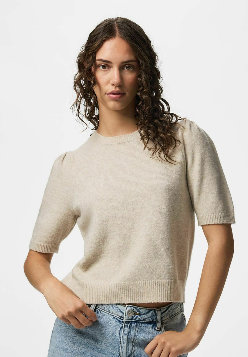 Femme aux cheveux bruns bouclés portant un pull beige à manches courtes et un jean bleu clair, posant devant un fond uni.
