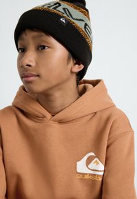 Bruin hoodie met een wit en geel logo aan de voorkant. Draag het met een zwarte gebreide beanie met oranje en teal patronen.