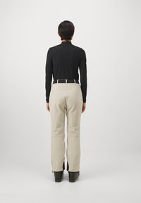 Schwarzes Langarmoberteil und beigefarbene gestreifte Hosen mit taillierter Taille, ausgestattet mit einem schwarzen Gürtel. Die Hosen haben Reißverschlüsse an den Knöcheln.