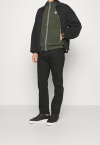 Svart zip-up jacka över en grön hoodie, svarta byxor och svarta sneakers. Har dragkedja och ränder på hoodien, avslappnad passform.