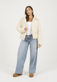 Doudoune beige côtelée avec un col montant et des boutons-pression. Associée à un jean bleu large et des bottines marron sur un fond neutre.