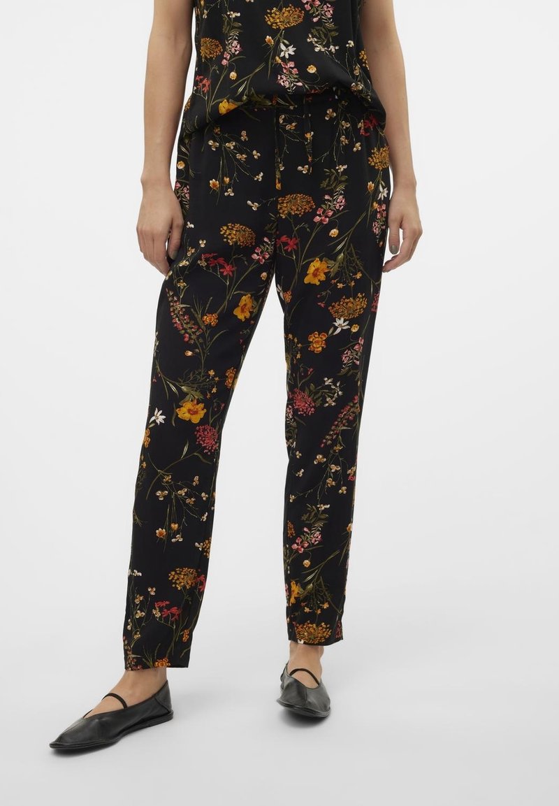Personne portant un pantalon noir fuselé avec un imprimé floral multicolore et des chaussures plates noires avec une bride sur le pied.